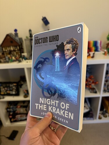 A hand holds a "Doctor Who: Night of the Kraken" book, with a bookshelf filled with toys and models in the background.