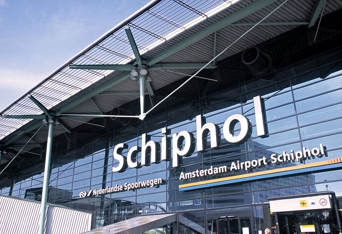 The image shows the exterior of Amsterdam Airport Schiphol, featuring prominent signage on a glass facade. (Captioned by AI)