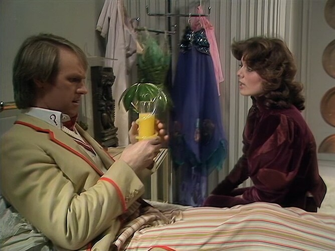 A man in a cream suit is sitting on a striped bed holding a yellow drink, while a woman in a burgundy outfit sits nearby in a room with hanging clothes. (Captioned by AI)