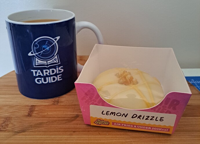 A "Lemon Drizzle" cake sits next to a mug labelled "Tardis Guide" on a wooden surface. (Captioned by AI)
