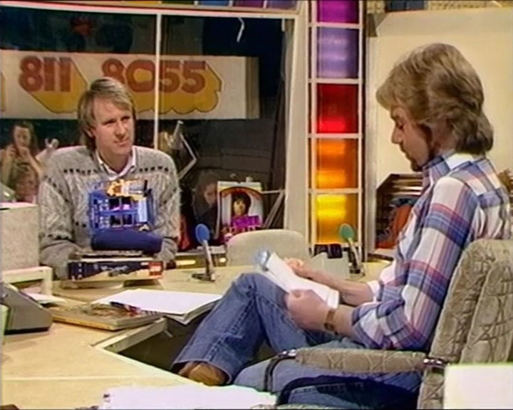 Two men are sitting in a brightly coloured studio, one holding papers and the other a toy, with a telephone number displayed in the background. (Captioned by AI)